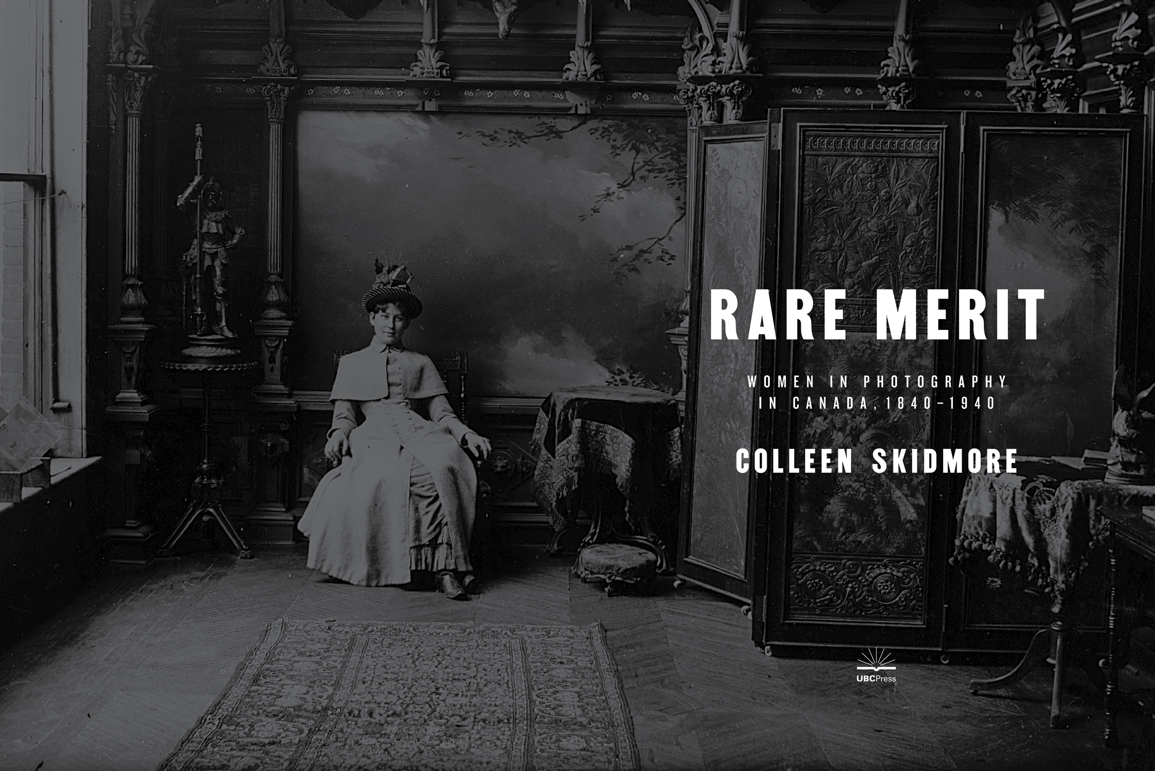 Title page image for ‘Rare Merit’ shows a black and white image of a woman sitting in an elaborately decorated room. The woman is wearing a dress, circa early 1900s, and a fancy hat.